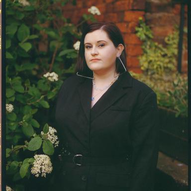 Szou wearing a black coat and triangle earrings, stood outside next to foliage